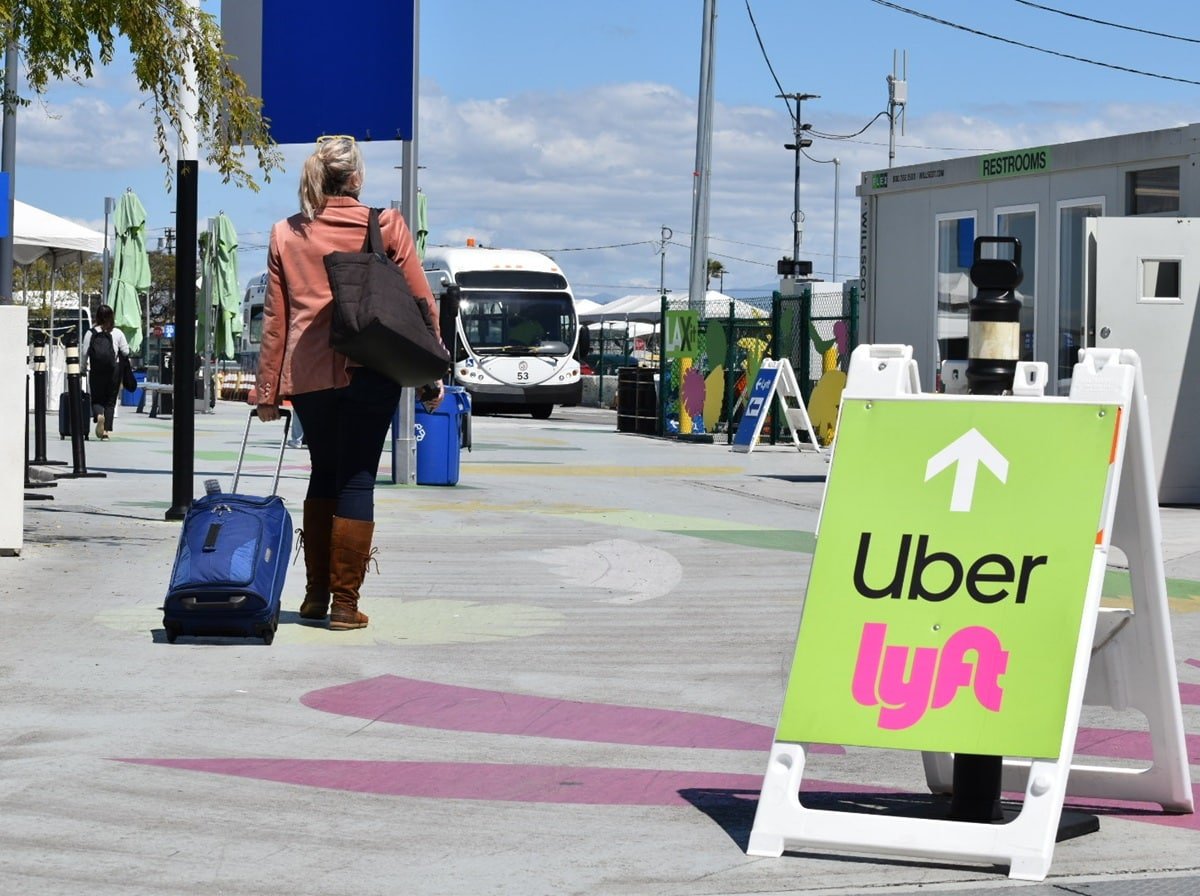 Photo of woman walking with luggage past a Uber/Lyft sign